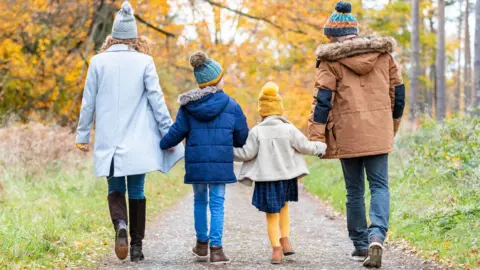 Getty Images A stock image of a family walking on a woodland path. On the right a woman wearing a winter coat, a bobbly hat and knee-high boots. Next, a boy wearing a blue park, a bobbly heat and jeans. He is holding hands with a young girl who is wearing a yellow hat and tights, with a coat and blue dress. On the right is a man wearing a hat and a brown parka jacket.