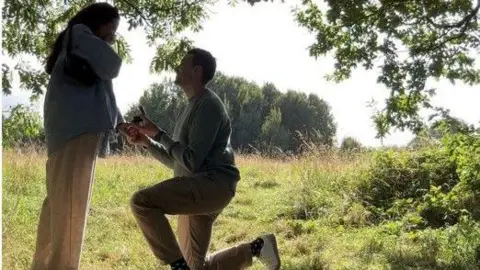 Sarah Burwin A woman holds her hands to her face in shock as a man gets down on one knee on the edge of a verdant wood on a sunny day. 
