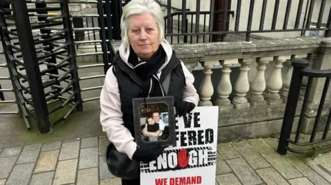 Martina Glenn, with grey hair and black and cream scarf, holding picture and placard