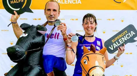 Jo Laplana A man and woman dressed in sports gear with inflatable horses around their waists. They are each holding medals in front of a backdrop which has the word "Valencia" printed on it repeatedly. The woman holds a sign in the shape of a speech bubble which reads "Supercity Valencia." The man is holding a similar sign which says "Halfs." 