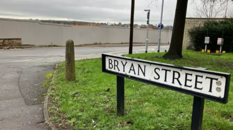 A large road sign in the foreground says Bryan Street, with a long grey wooden fence on the other side of the road.
