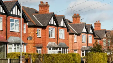 A row of two-storey, semi-detached houses on a residential street, with a hedgerow in front.