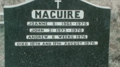 A gravestone with the nhame 'MAGUIRE' engraved in black capital letter into a surrounding block of white. The names 'JOANNE', 'JOHN' AND 'ANDREW' are also engraved in a similar way. At the top is a cross and beyond the white is granite. 