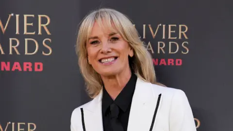 Tamzin Outhwaite smiling for the cameras in a white blazer. She is stood beside a photo board with the logo of the Olivier Awards.