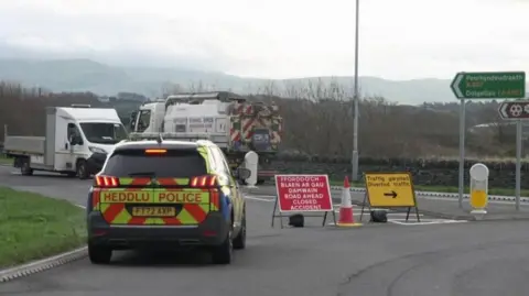a police vehicle blocks a road next to a sign that reads: 'Road ahead closed accident' and a traffic cone. Two large vehicles can be seen on the opposite side of the road.