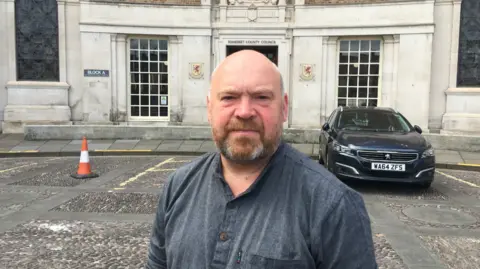 Daniel Mumby Bill Revans, in a grey casual shirt standing outside county hall in Taunton with a large black car just behind him