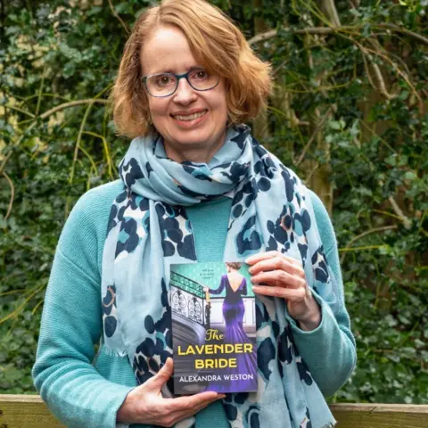 Tim Pearson A woman with short ginger hair and blue-rimmed glsses wearing a blue dotted scarf, turquoise jumper and holding a book called The Lavender Bride which shows a woman walking up a stair in a purple gown. A bush can be seen behind her.
