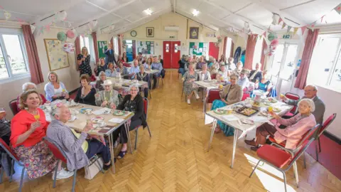 Re-engage A large village-hall type room with trestle tables and chairs laid out, all with a group of people sat round with tea and cake. All have turned to look at the camera, smartly dressed. There is bunting and balloons.