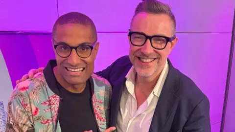 BBC Richie Anderson e Ed James parecidos um ao outro. Anderson, à esquerda, está usando óculos e uma jaqueta colorida sobre uma camiseta preta. James também está vestindo óculos de estrutura escura e uma jaqueta escura sobre uma camisa branca