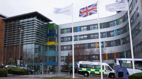 A shot of Queen Alexandra Hospital, a large building with lots of big glass windows and coloured panels. An ambulance and a bus can be seen outside. Three flags, including a Union Flag, are flying outside the building.