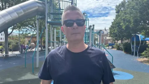 Jeff is wearing a dark T-shirt and sunglasses and is standing in front of a slide at a playground.