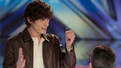 Chris Haston/NBC Magician Joel Mawhinney holds up a small silver key in one hand while talking to the judges on the America's Got Talent TV show. He has  short, curly brown hair and is wearing a brown leather jacket and a white shirt.  He is facing Simon Cowell who has his back to the camera.