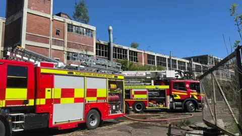 Two fire engines in bright red and yellow colours are parked outside a derelict old brick factory with a blue sky overhead