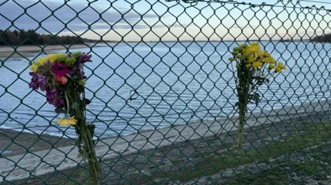 Floral bouquet that has been strapped to a metal fence. They are made of flowers that are different shades of yellow, orange, pink and red. A reservoir can be seen on the other side of the fence. 