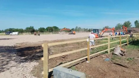 SOUTH CAMBRIDGESHIRE DISTRICT COUNCIL Diggers working on the site, where hard standing has been put down. There are several people standing on the site. A three-barred wooden fence is in the foreground, along with the detached bucket of a digger.