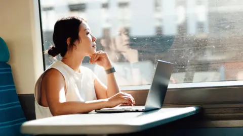 Getty Images Young business woman looks out the train window while using her laptop on a table