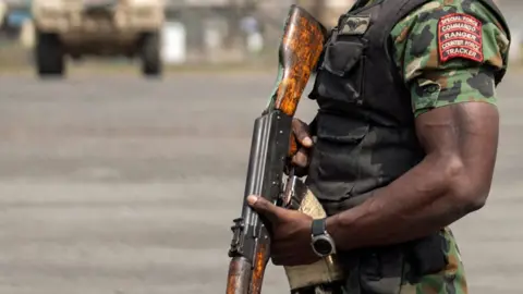 A uniformed Nigerian soldier holding a gun with donated US armoured trucks in the background