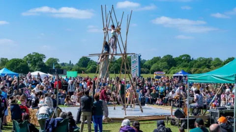 An acrobatic performance of two people standing on bamboo sticks with people watching them. 