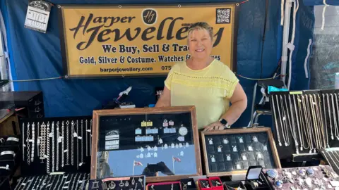 Darren Rozier/BBC A woman stands behind a jewellery market stall. She is smiling and is wearing a yellow top. She has blonde hair that has been tied back. Various necklaces and other pieces of jewellery are on display in cases in front of her. 