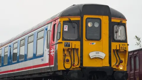 The Clacton Express pictured after the restoration works on its exterior. The electric train has a yellow front and a sign that reads: "309 Homecoming." On its side it is painted blue with white and red detailing. A crane can be seen behind the vehicle.