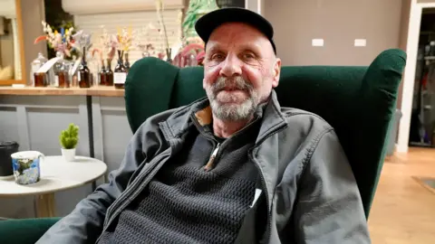 David Rain is wearing a black hat and sitting in a green arm chair in the coffee shop in the market.