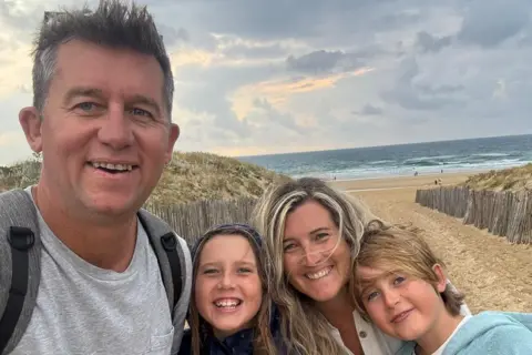Zack Van Aarde A family of four are smiling into the camera while standing on a beach. The dad is closest to the lens, while the daughter and son are leaning into their mum. 