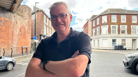 Andy Woodger smiles into the camera with his arms crossed while standing on the pavement with the kerb. The mini roundabout with cars is behind him. He is wearing glasses and a black polo top