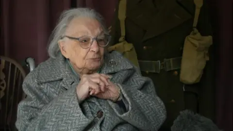 A woman wearing a grey coat sits with her hands clasped together.