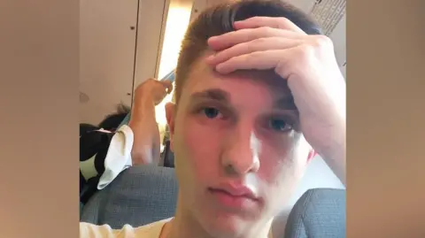 SamWilliams Sam Williams sat on a train with his hand pressed against his forehead looking disappointed