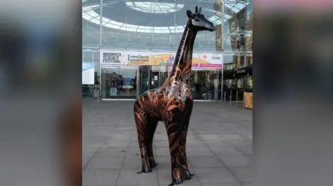 Break A statue of a giraffe, which is painted black and gold, is standing on a concrete walk way. In the background is a large building made of glass, with an entrance at the front.