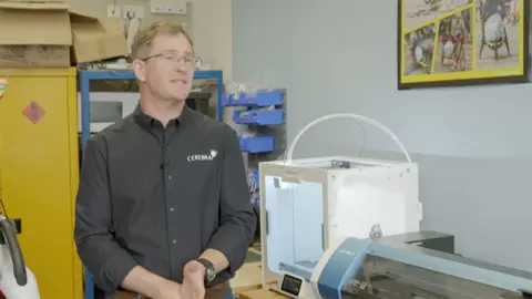 Dr Ross Head from Cerebra he has brown hair and wears glasses and is wearing a black shirt with the organisation name in white letters on the left breast he is standing in front of a 3D printer and a cutting machine