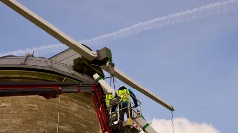 Men in a cherry picker working on windmill stocks with a vapour trail above in a blue sky