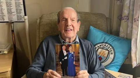 John Cunningham sitting on a chair smiling with resting his back on a  Manchester City FC cushion holding up a 100th birthday card showing King Charles and Queen Camilla 