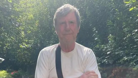 Leslie Midwinder, a man with short hair brushed to the side, is pictured looking at the camera. He is stood outside in front of some green trees and bushes and wearing a white T-shirt.