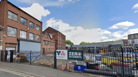 Google A large red brick building is on the left with a small plaque that reads Banks Brewery. There is a huge yard at the front with hundreds of metal beer kegs piled on top of each other, behind a black metal fence.