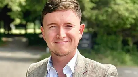 John Birtles John Birtles, who has dark blonde hair and light stubble on his chin, smiles at the camera wearing a gold coloured blazer over a white open necked shirt. 