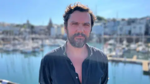 Oliver is wearing a blue shirt and has short dark brown hair as well as a beard. He is standing at the harbour in St Peter Port in Guernsey.