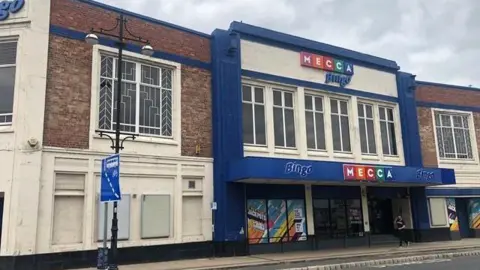 A 1930s Art Deco-style brick building with white and blue moulding. On the front are signs for Mecca Bingo.