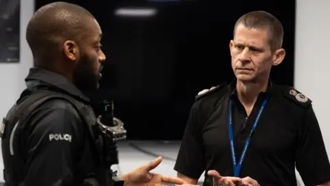 Chief Constable Ivan Balhatchet talking to a police officer, who is to his left. He looks serious, with a blue lanyard round his neck, wearing a black police top. The other officer, is looking at him. They are both standing in a room. 