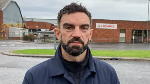 A man with dark hair and a dark beard looking at the camera with a serious expression on his face. He is wearing dark outdoors clothing and standing by the side of the road in front of the main entrance, a red brick building, for Leonardo Yeovil which has a sign reading 'Leonardo' with the company logo