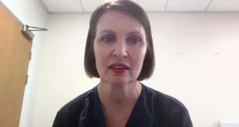 Griffin sitting indoors. She is wearing a dark blue top, and her hair is cut into a neat, chin‑length style. The background features a plain light‑coloured wall, a closed wooden door with metal hinges and a handle, and a white ceiling with recessed lighting panels.