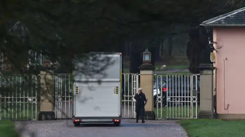 Getty Images A white van parked up at white gates, where a person stands, speaking into a radio. The shadow of a tree obsecures the view in the foreground.