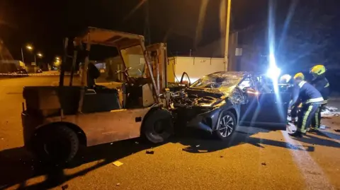 Three firefighters are working next to a badly smashed up car with open driver and passenger door, missing bonnet and wrecked windscreen that is sat in front of a forklift truck. 