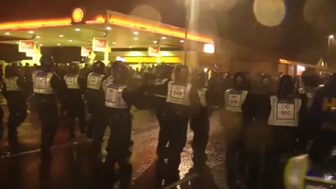Helmeted police officers in a line confront fans at night in a street outside a petrol station.