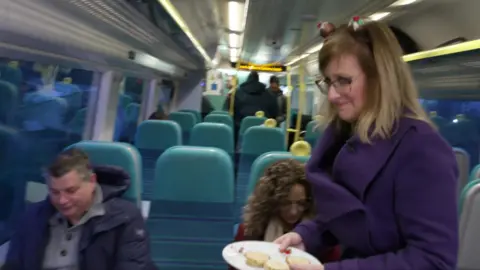 A woman wearing a purple coat and a Christmas party hat tries unsuccessfuly to hand out mince pies from a plate aboard a Southern Railway commuter train.