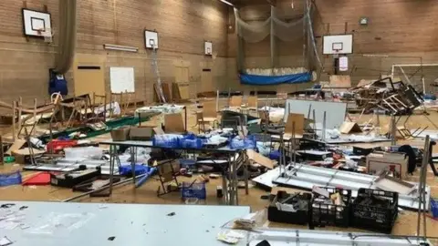 Market Deeping Model Railway Club A sports hall with chairs and tables in it which has been turned upside down and vandalised. There are items scattered all over the place.