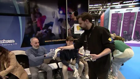 A man with grey and brown hair and facial hair in a blue top is sitting down on a bench and reaching out to a man handing him a large pair of white gloves. The man with the gloves has brown hair and facial hair, a black jumper and a black lanyard on 