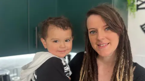 A woman is holding her two-year-old son to the right of her. They are both looking directly into the camera smiling. They are standing in the kitchen in front of worktops. 