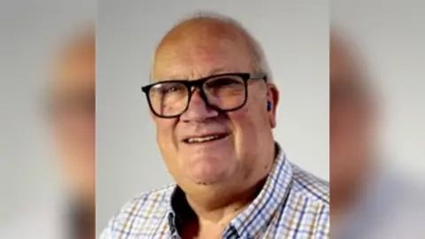 Rob Howard, a man wearing a checked shirt and glasses, is stood against a light-coloured backdrop.