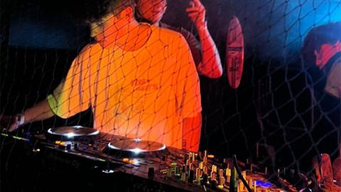 Shelly DJing. Shelly is an 18-year-old man with short brown hair. He wears an oversized white T-shirt and is pictured in a dark club lit by an orange light as he stands behind some decks. 